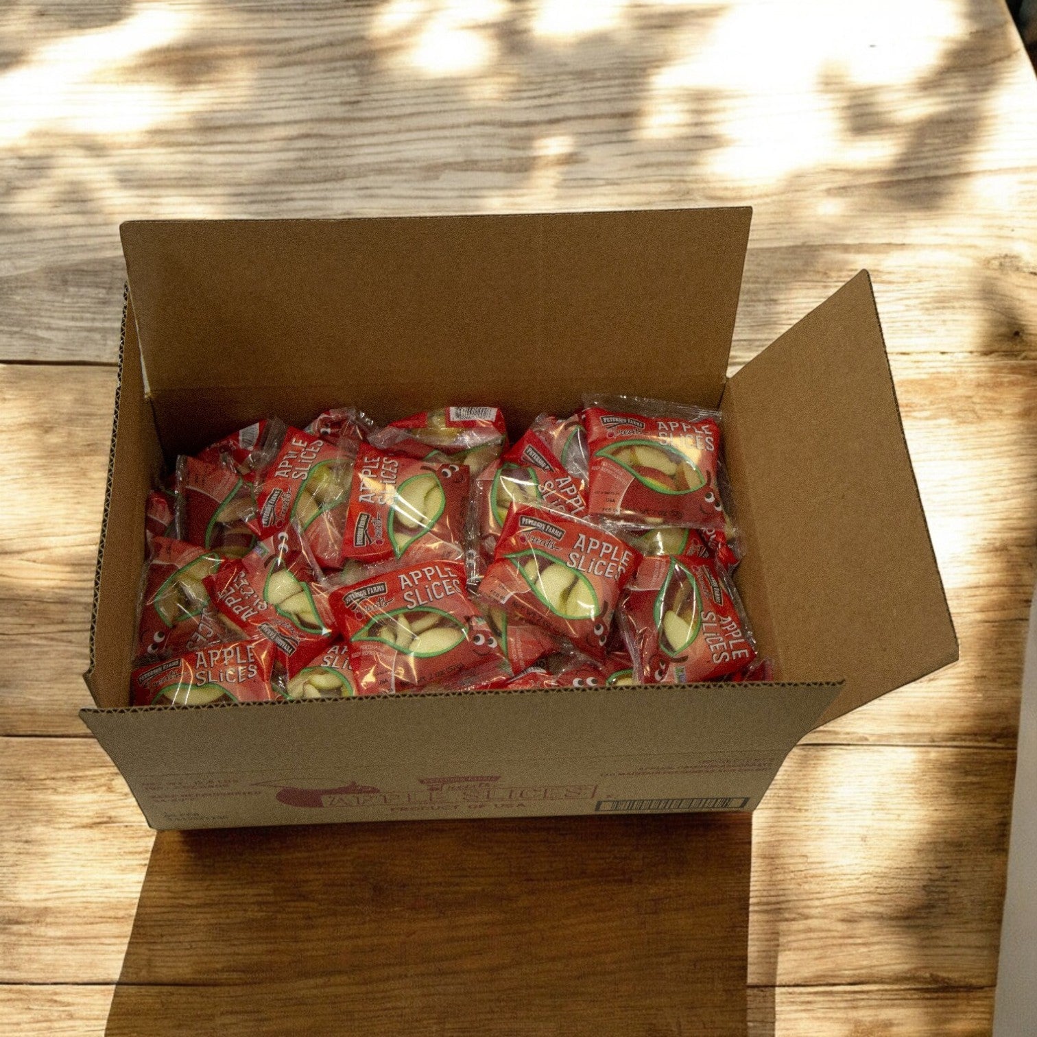 A Peterson Farms cardboard box containing Fresh Sliced Apple Packs for Convenient Snacking (2 oz., 1 case, bulk 100) sits on a wooden surface, contrasted by light and shadow patterns.