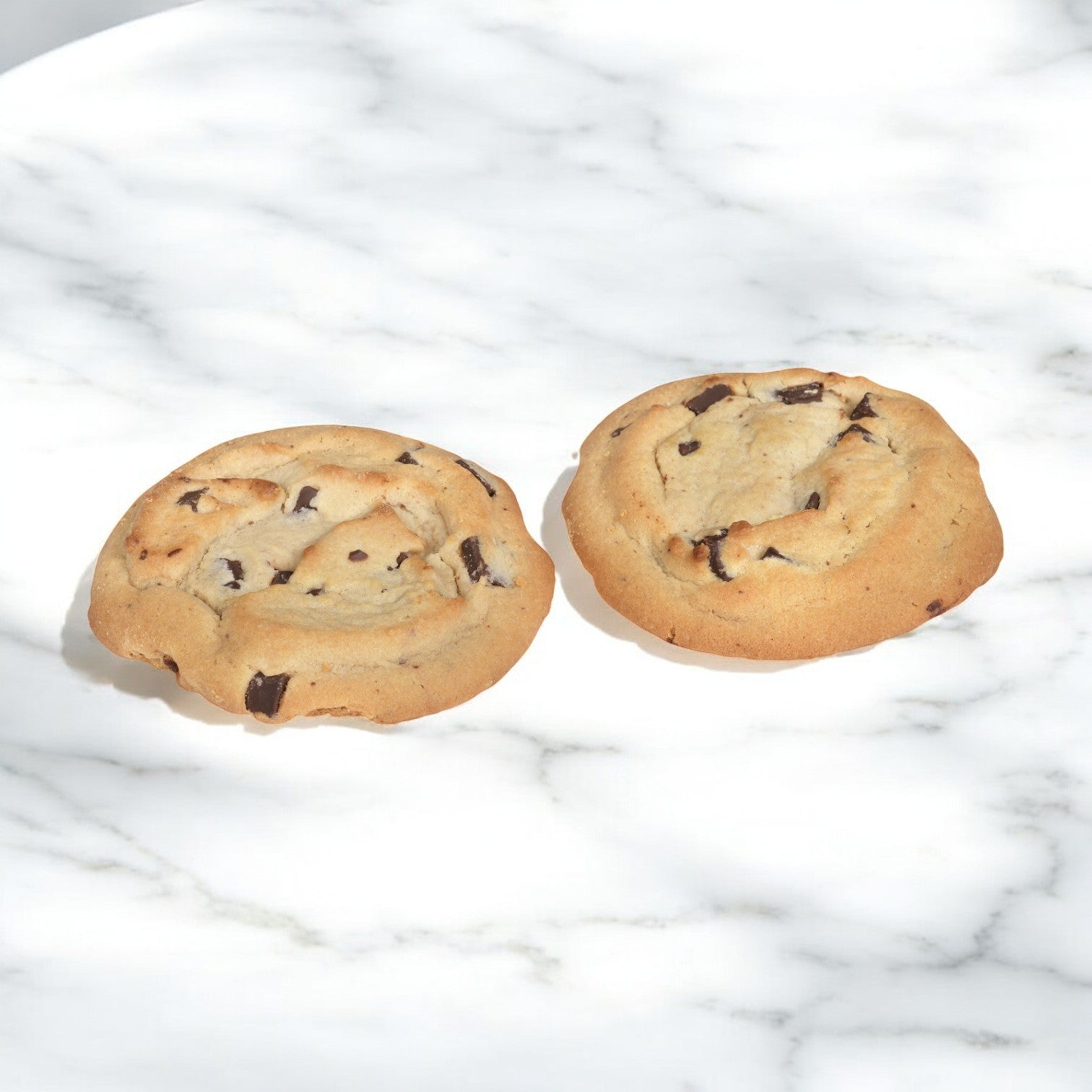 Two Otis Spunkmeyer Cookie Chocolate Chunk Bulk Thaw & Serve cookies rest on a white marble surface, partially lit by sunlight.