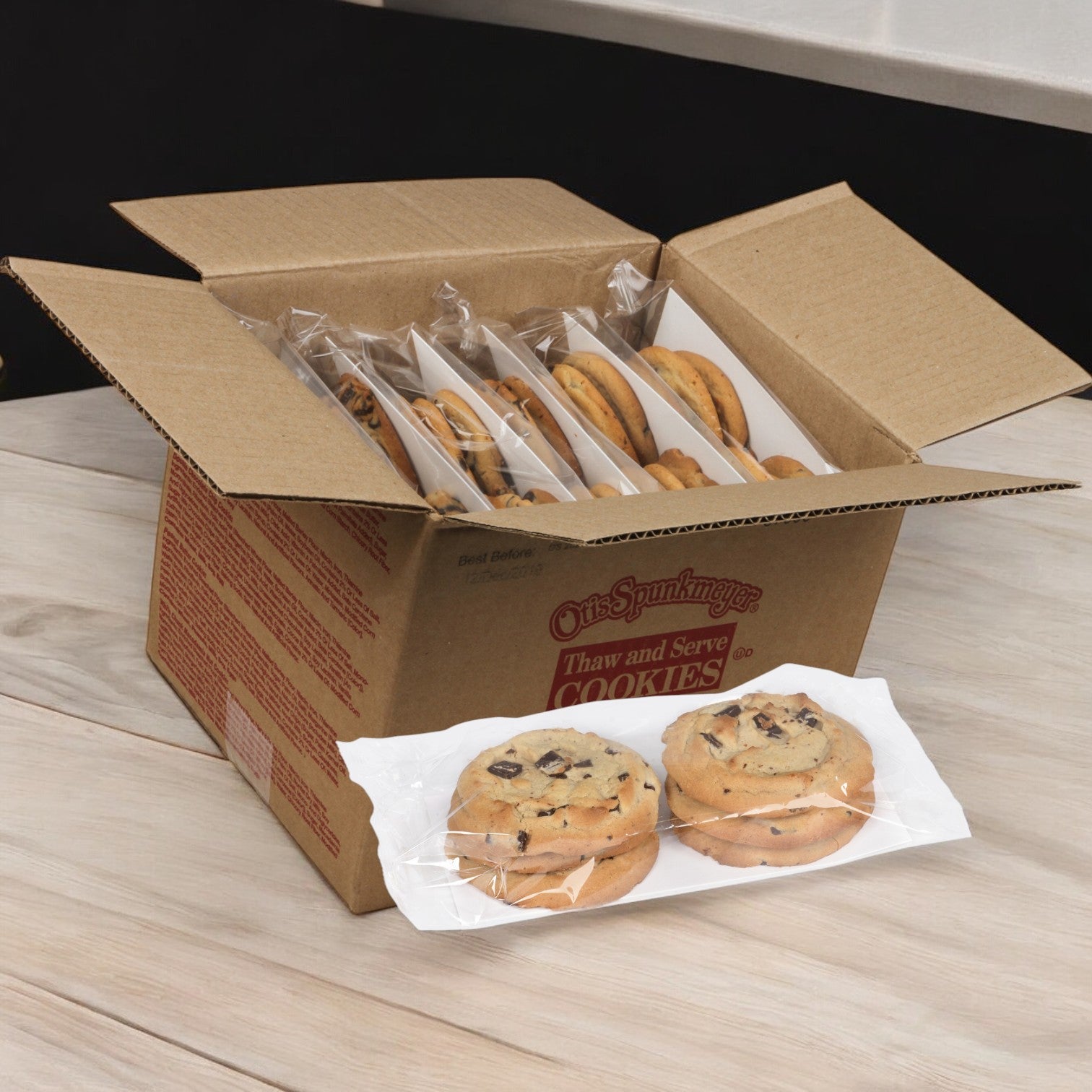 A case of Otis Spunkmeyer Cookie Chocolate Chunk Bulk Thaw & Serve containing 72 cookies is on a wooden table, featuring rich semi-sweet chocolate chunks ready to delight your taste buds.