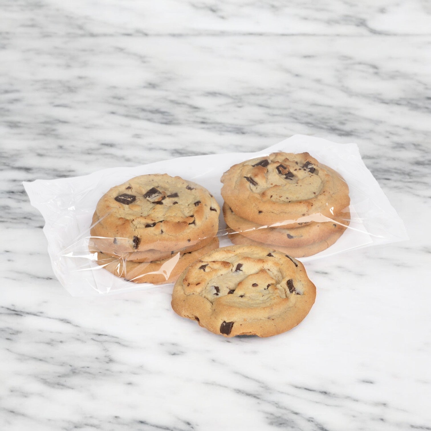 Three Otis Spunkmeyer Cookie Chocolate Chunk cookies lie invitingly on a marble surface, wrapped in clear plastic, with tempting semi-sweet chocolate chunks. Perfect for a delightful thaw and serve moment, part of a case offering 72 treats from the brand Otis Spunkmeyer.