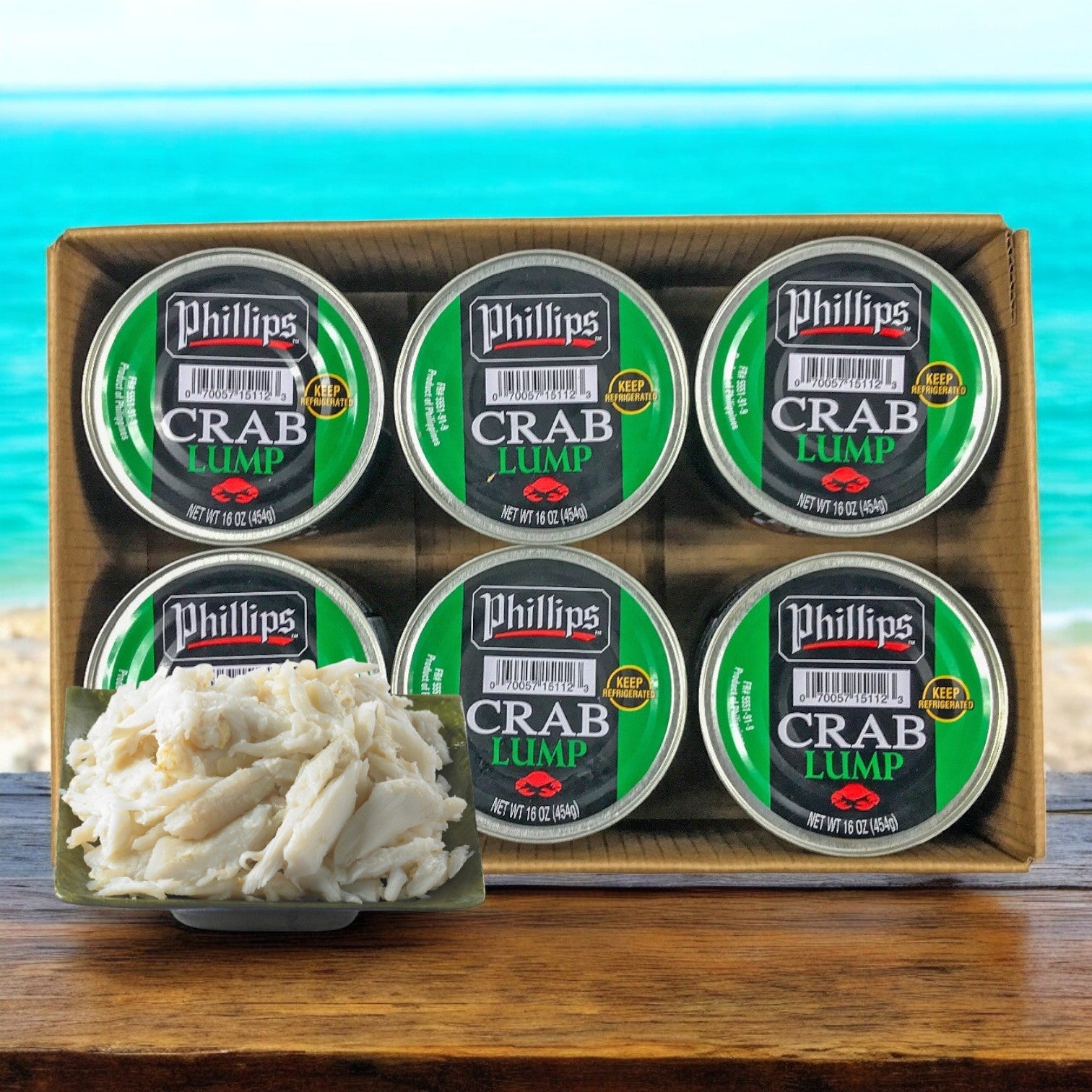 A case of Phillips Foods Crab Meat Lump Pasteurized, featuring six 1 lb. cans, sits on a wooden surface. A small plate displays jumbo lump crab meat, with a turquoise sea and sandy beach in the background, inspiring versatile culinary use.