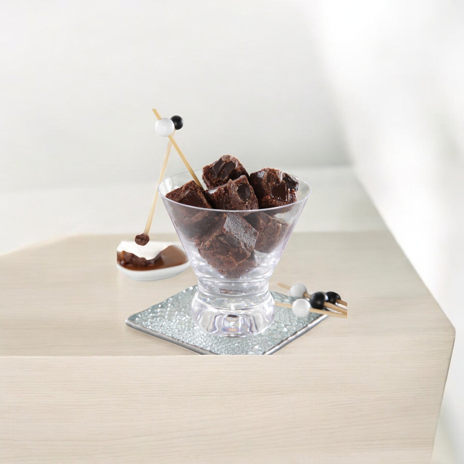 A glass cup filled with Sweet Street Brownie Chocolate Bites from a 121-count case sits on a silver coaster, with decorative skewers beside it on a white surface.