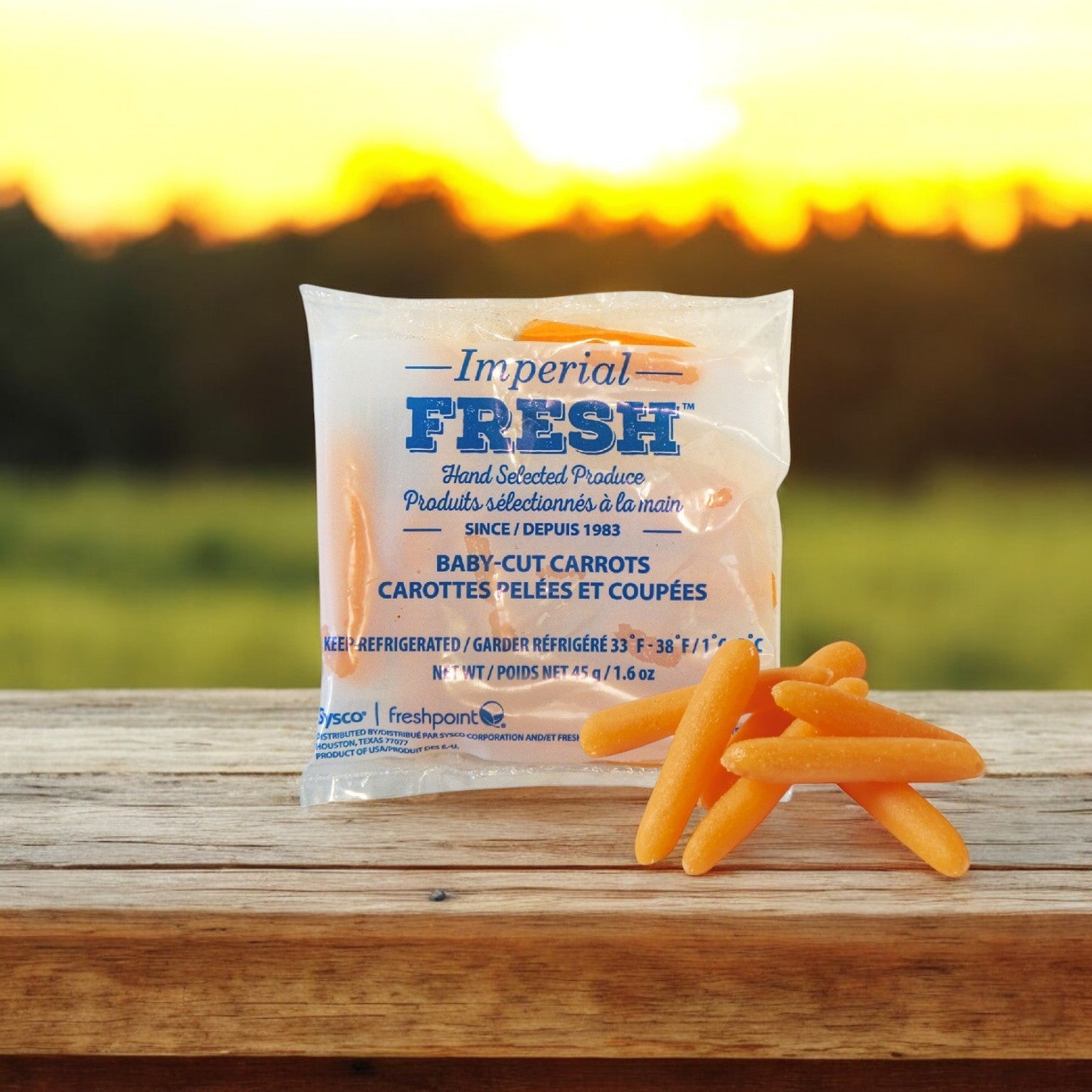 A bag of Sysco Baby Carrots in Bags—Cut Peeled 1.6 oz. (200 Bags) sits on a wooden surface, loose carrots nearby; fields and a sunset form an inviting backdrop for this fresh, healthy snack.