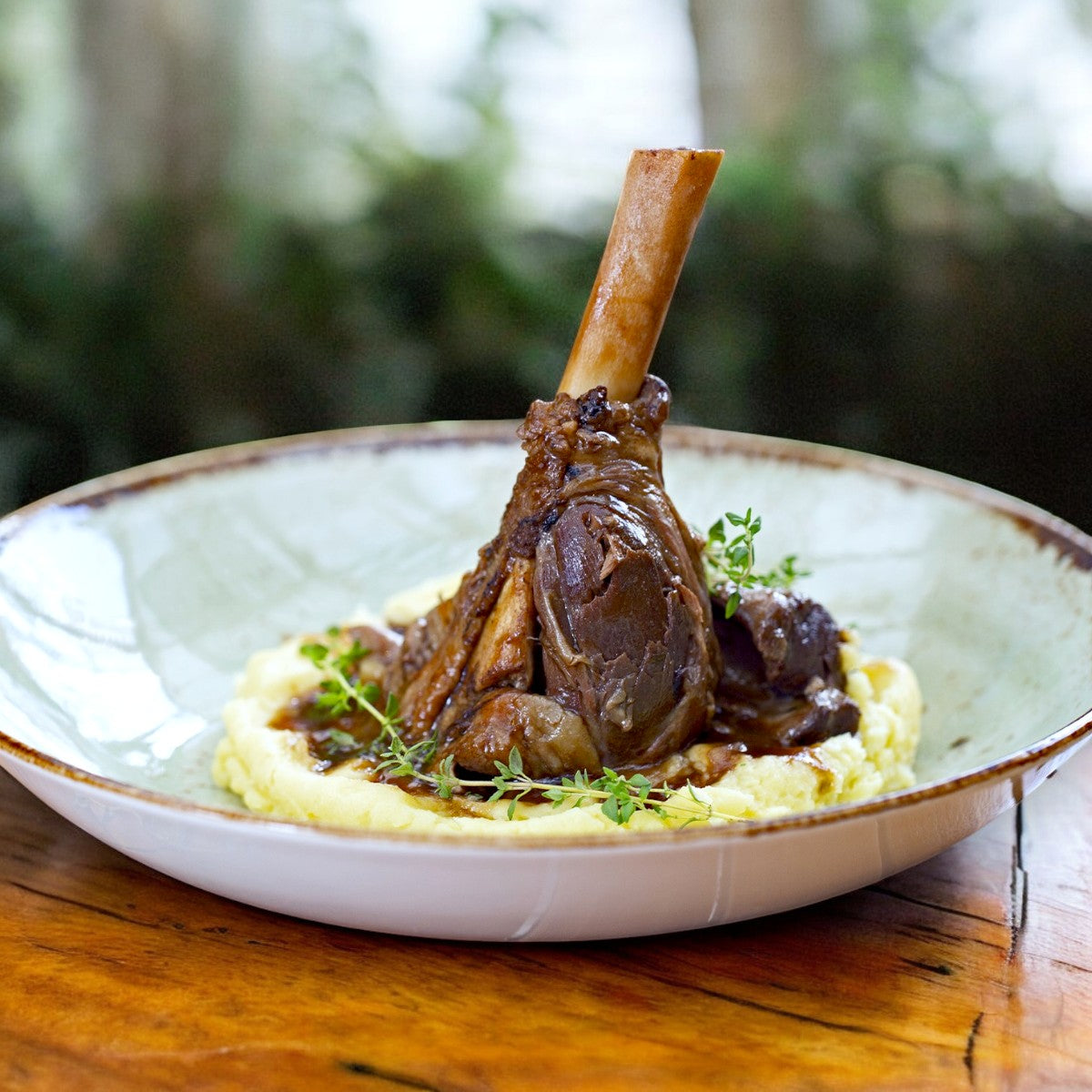 The Butchers Block by Sysco Halal Lamb Hindshank, from the 20-pack (16 oz each), is served on mashed potatoes and garnished with fresh herbs in a shallow bowl on a wooden table.