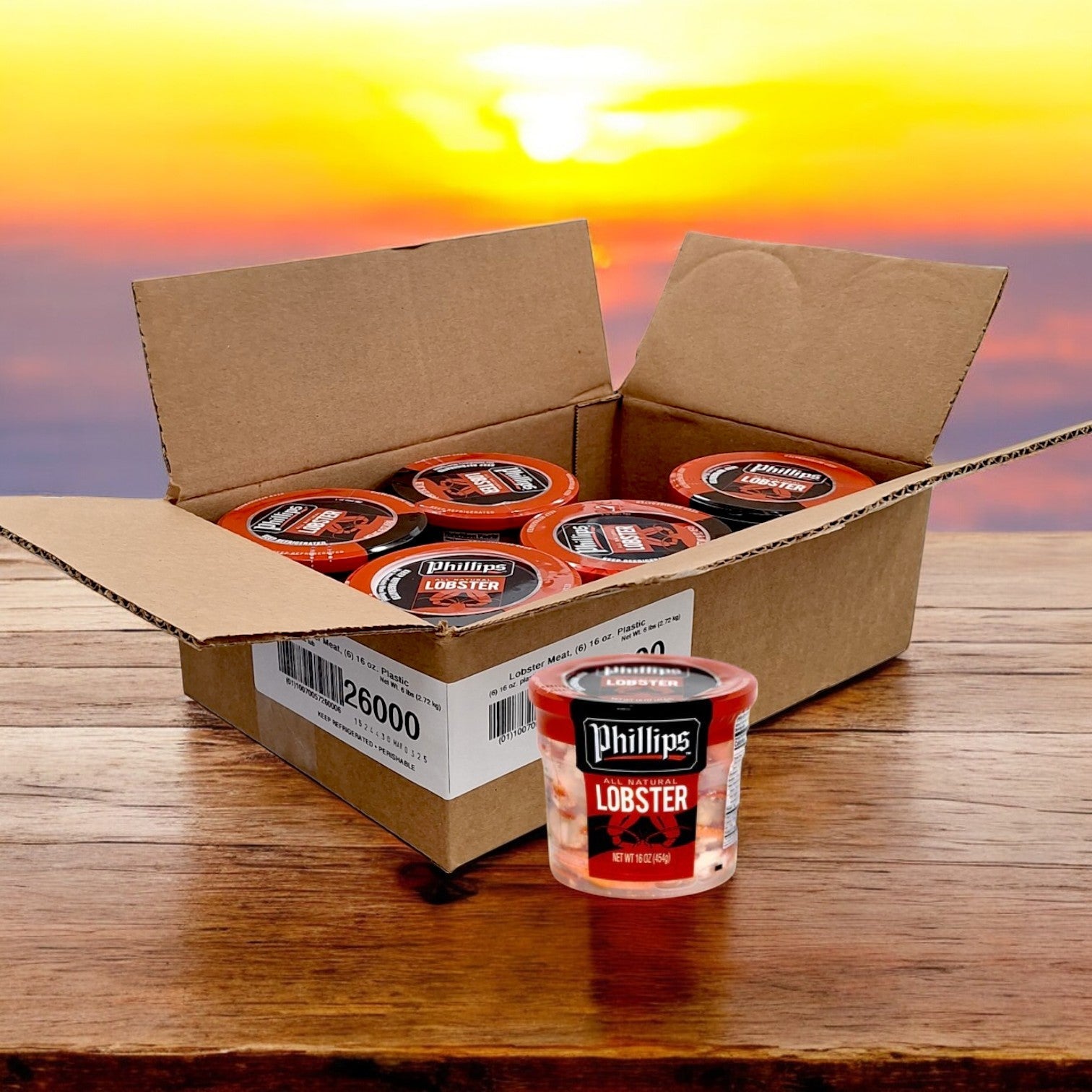 An open cardboard box displays several containers of premium seafood, including a package of Phillips Foods Lobster Meat – 16 Ounces (6/16 oz) placed outside. The background features a stunning sunset view, highlighting this ready-to-use delicacy.
