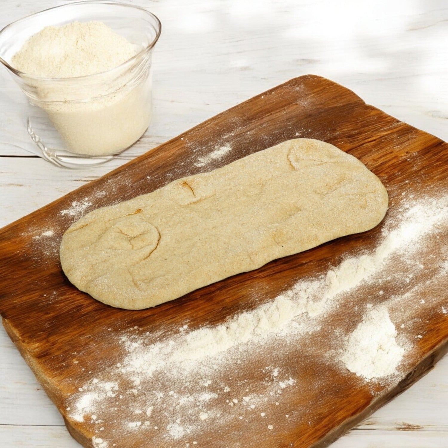 Flat oval dough from Rich's, sized 12" x 5", rests on a wooden board lightly dusted with flour, ready to be made into fresh-baked flatbreads, alongside a glass bowl of flour.