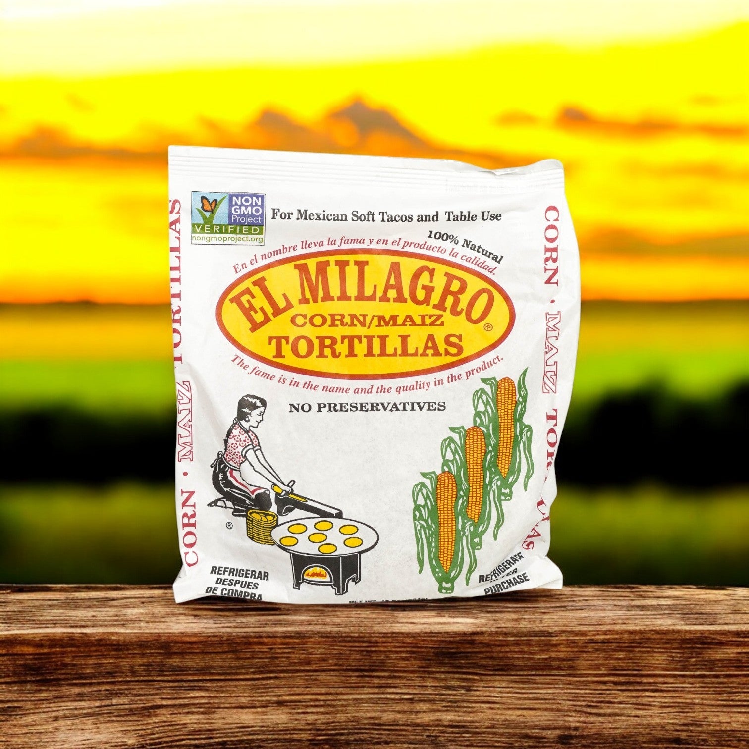A package of El Milagro Corn Tortillas, Yellow 6" One Dozen captures traditional Mexican cuisine on a wooden surface, with a vibrant field under a colorful sunset in the background.