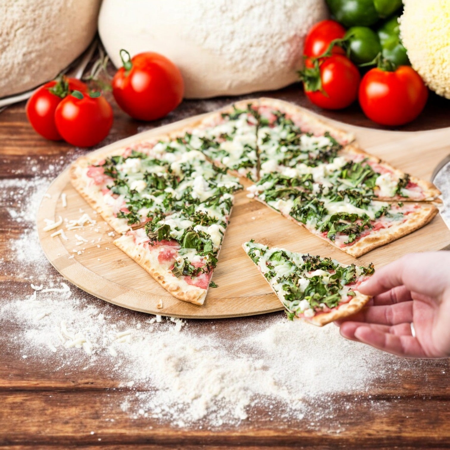 A hand picks up a slice of Rich's Bread Flat Thin Oven Fired pizza topped with greens and cheese on a wooden board, surrounded by tomatoes, dough, and green peppers on a floured surface.