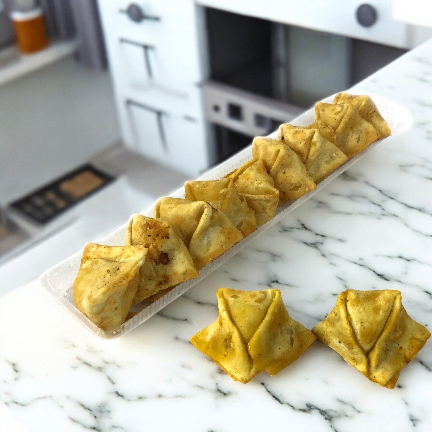 A tray of triangular Crab Rangoon pastries made with wonton wrappers and visible spices rests on a marble counter, with two extras nearby. Blurry kitchen appliances in the background enhance the charm of a kitchen preparing Sysco's frozen appetizers from a 100 count case.
