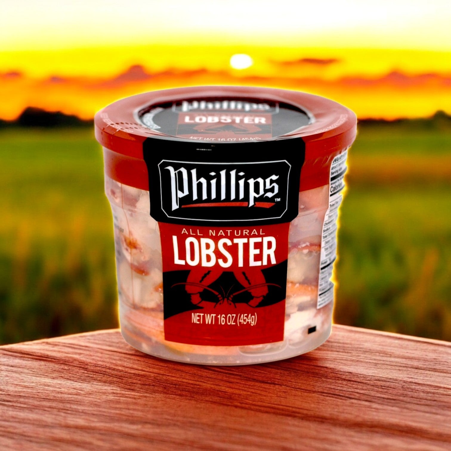 A container of Phillips Foods Lobster Meat, 16 oz, rests on a wooden surface at sunset over a grassy field. This pasteurized treat is ideal for gourmet seafood dishes.