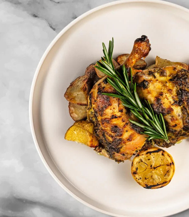 Savor the FoodHaul Roasted Half Brick Chicken, boasting crispy skin and garnished with sliced lemon, potatoes, and a sprig of rosemary on top.