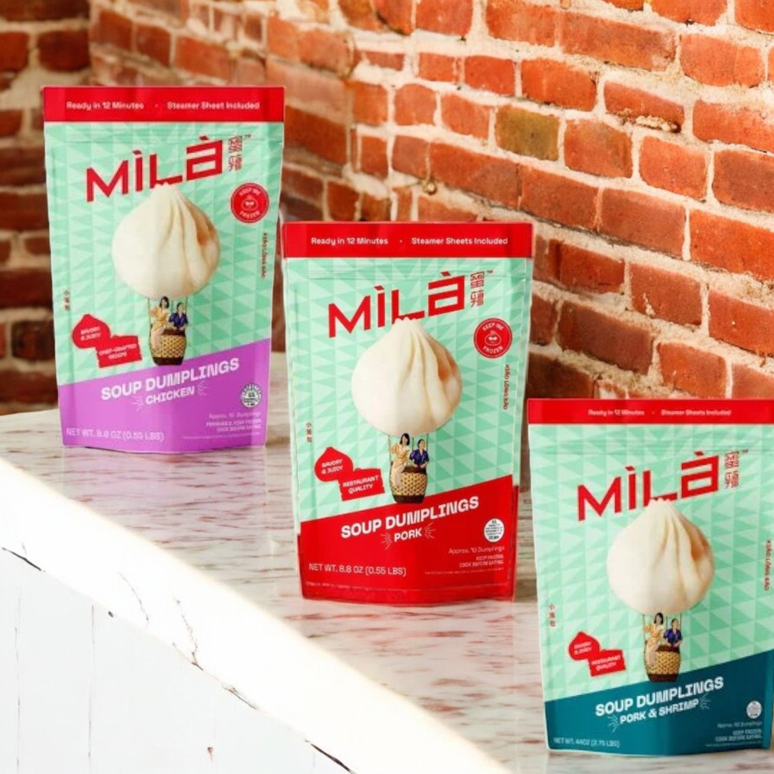The Mila 3 Pack Soup Dumpling Combo rests on a white surface against a brick wall, featuring authentic flavors of chicken, pork, and pork & shrimp.