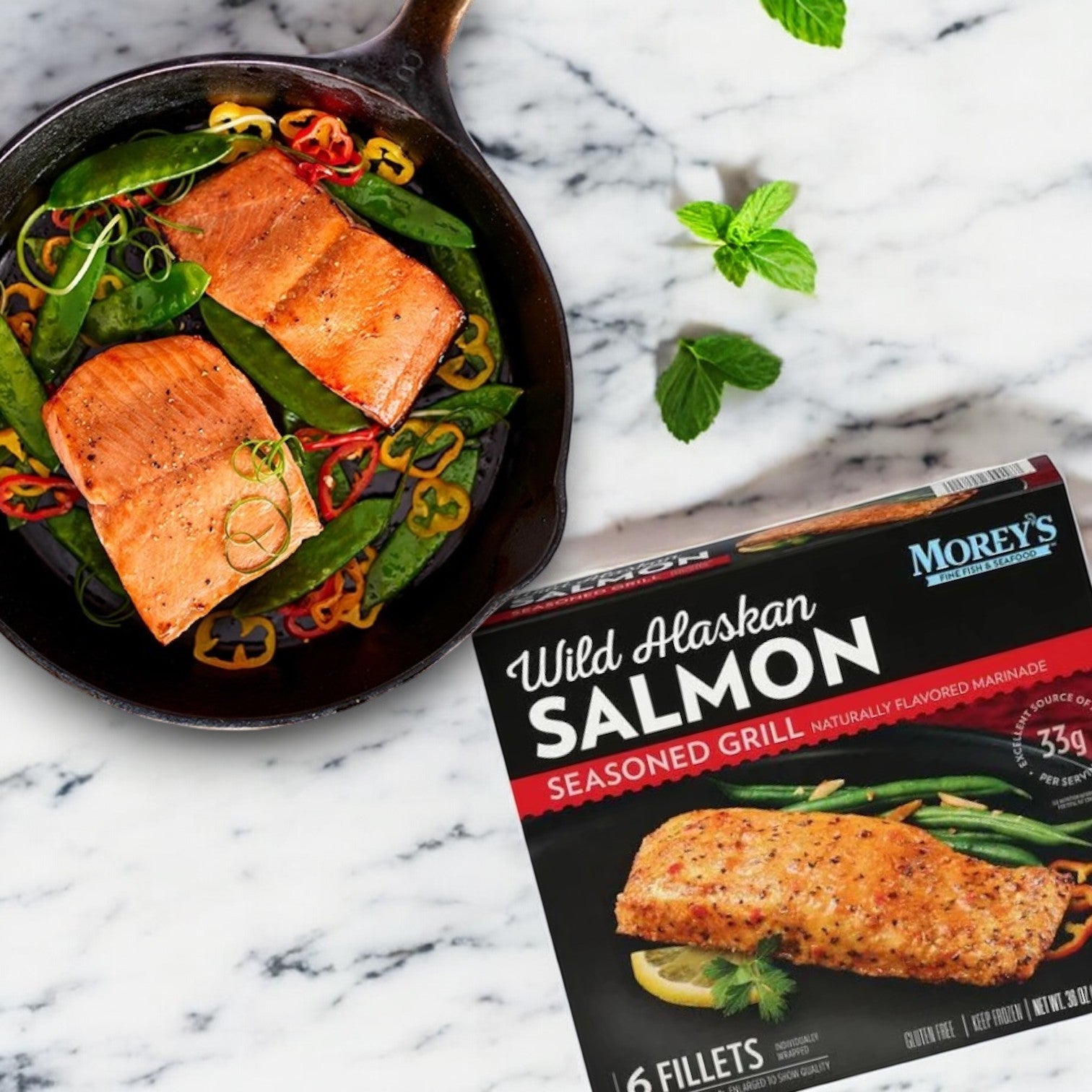 A cast iron pan with cooked wild-caught salmon, snap peas, and sliced peppers sits next to a box of Morey's Fine Fish & Seafood Marinated Wild Alaskan Salmon, 6 ct-1 Box on a marble surface.