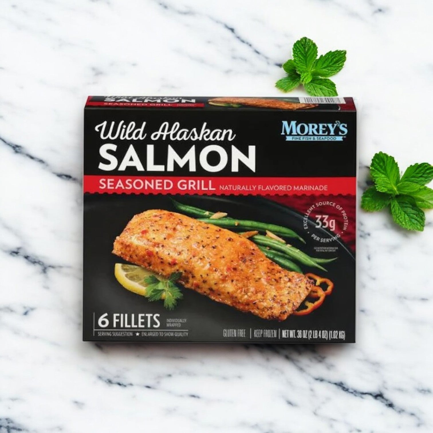 A box of Morey's Fine Fish & Seafood Marinated Wild Alaskan Salmon, 6 ct, rests on a white surface with two sprigs of fresh mint beside it.