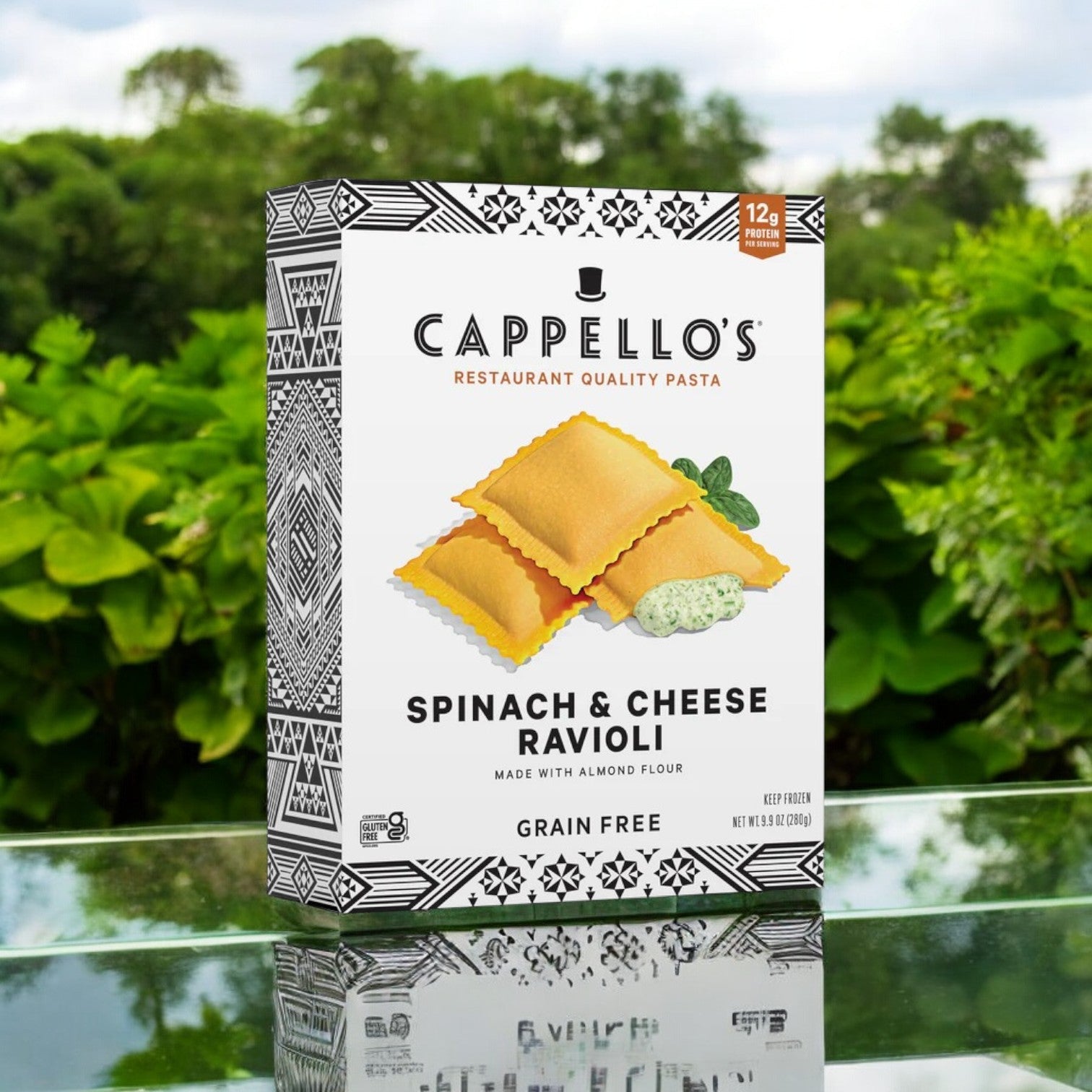 A 9.9 oz. box of Cappello's Spinach & Cheese Ravioli with almond flour pasta is showcased on a reflective surface outdoors, surrounded by lush greenery.