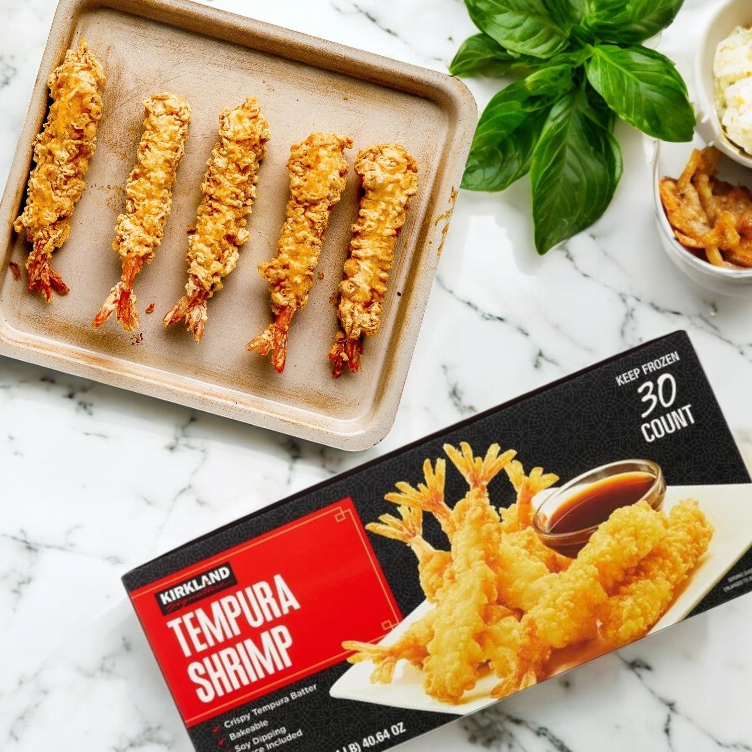 Five crispy Kirkland Signature Tempura Shrimp are arranged on a baking sheet next to a 2.35 lb, 30-count box, fresh basil, and a small bowl of sauce on a marble surface.