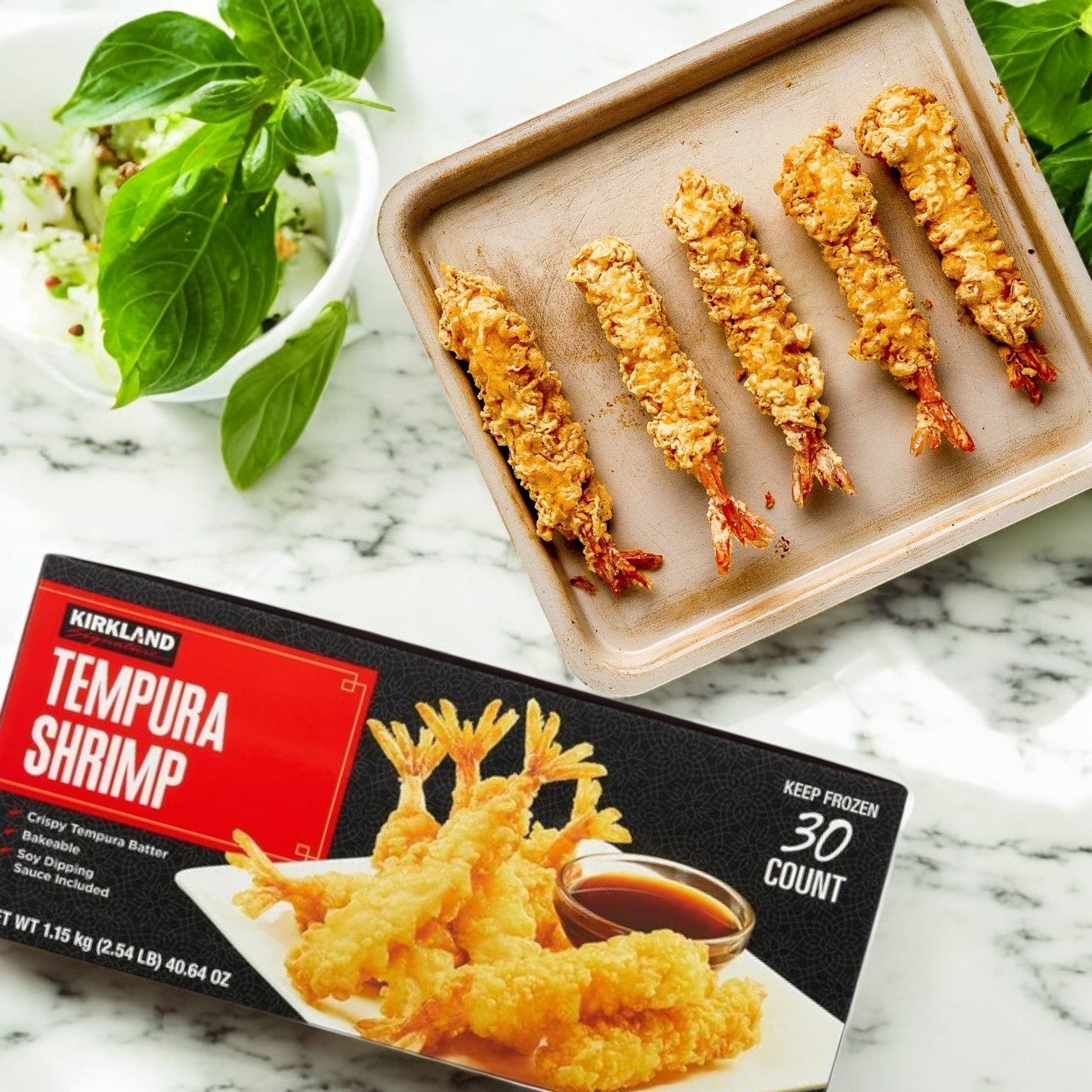 Five Kirkland Signature Tempura Shrimp sit on a tray beside a box labeled "Tempura Shrimp, 2.35 lbs, 30-count - 3 Packs" by Kirkland Signature, on a marble countertop with fresh basil in the background.