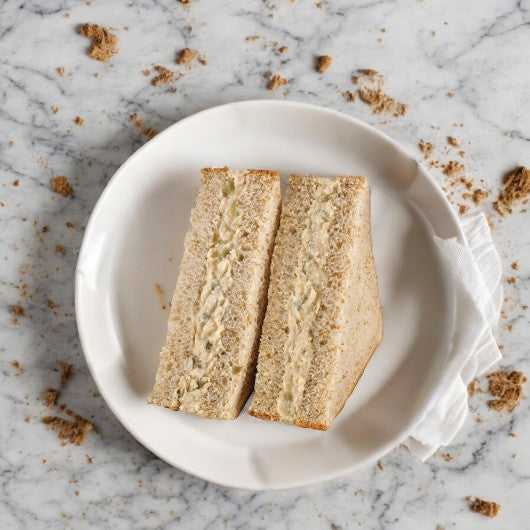 Two triangular halves of a creamy Albacore Tuna Salad Sandwich from E.A. Sween rest on a white plate atop a marble surface with scattered crumbs.