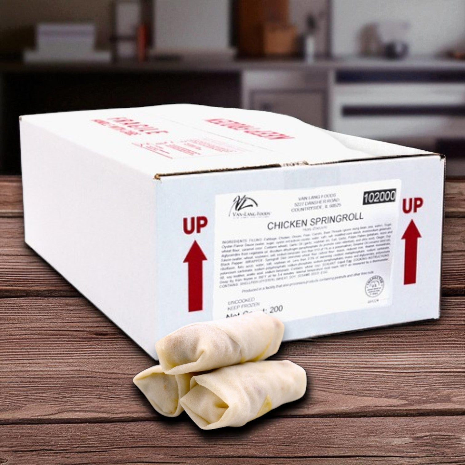 A Van Lang Chicken Spring Rolls – 200 Count (2 trays of 100, frozen appetizers) box sits on a wooden surface with three uncooked spring rolls in front; an ideal Asian appetizer, kitchen background visible.