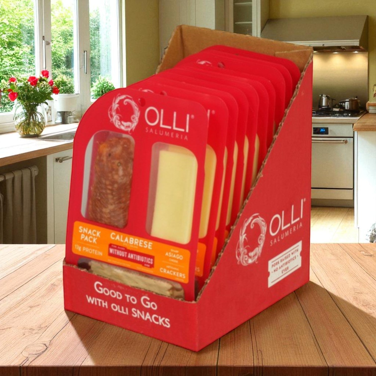 A red display box of Olli Salumeria Calabrese Salami & Asiago Cheese Snack Packs (10 pack, 2 oz each) sits on a wooden kitchen table, featuring high-protein snacks with salami, Asiago cheese, and crackers.