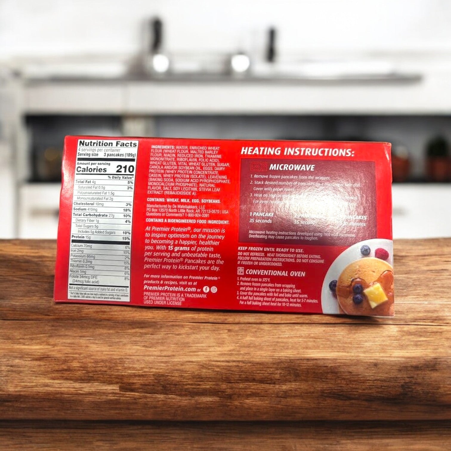 A box of Premier Protein Pancakes – 12 Count- 1 Pack from Premier Protein is placed on a wooden surface against a vivid red backdrop, highlighting its high-protein and nutritional benefits. Visible heating instructions complement the blurred kitchen background for added depth.
