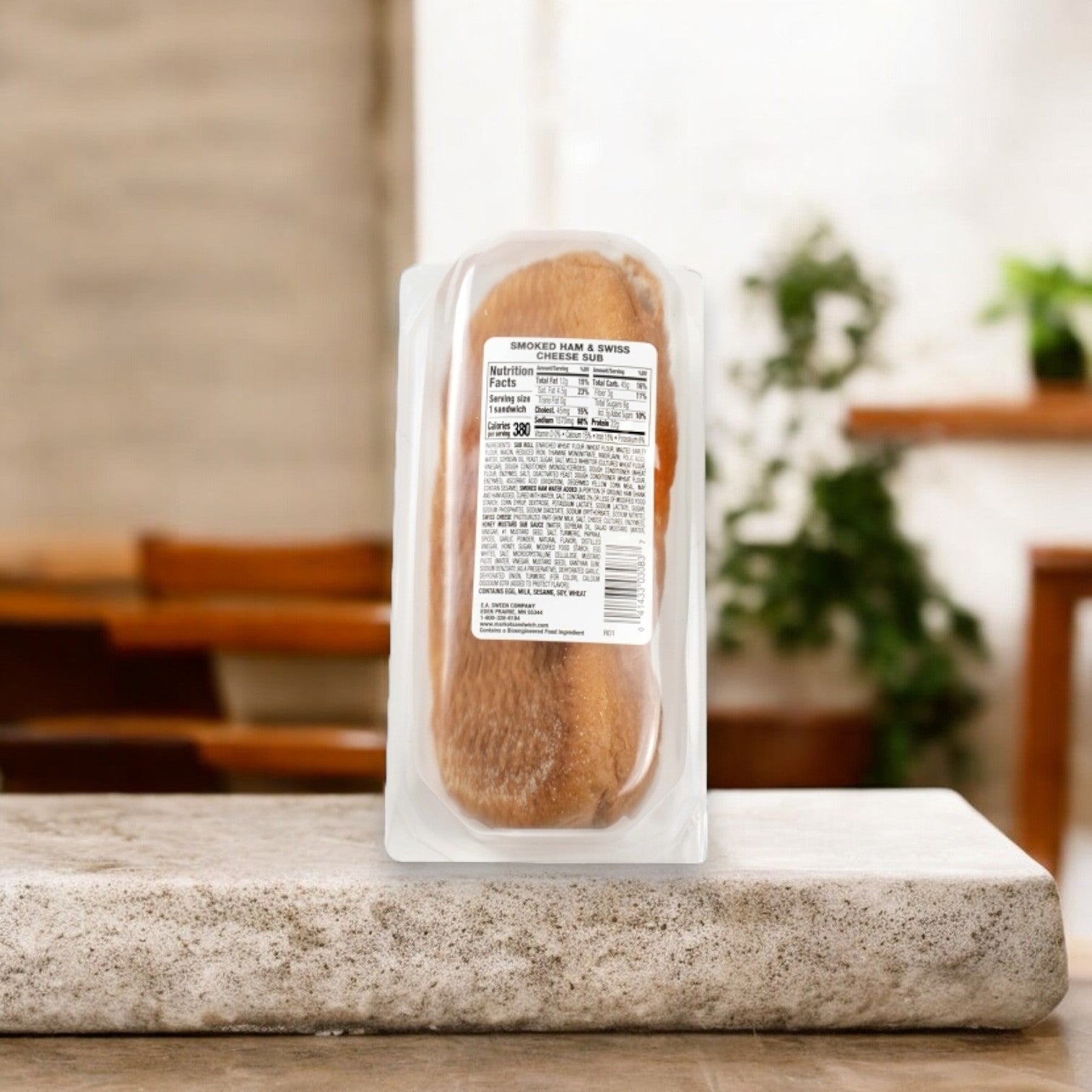 A Smoked Ham & Swiss Cheese Sub by E.A. Sween rests on a stone counter, showcasing its nutrition label amid tables and plants that enhance the calm ambiance.