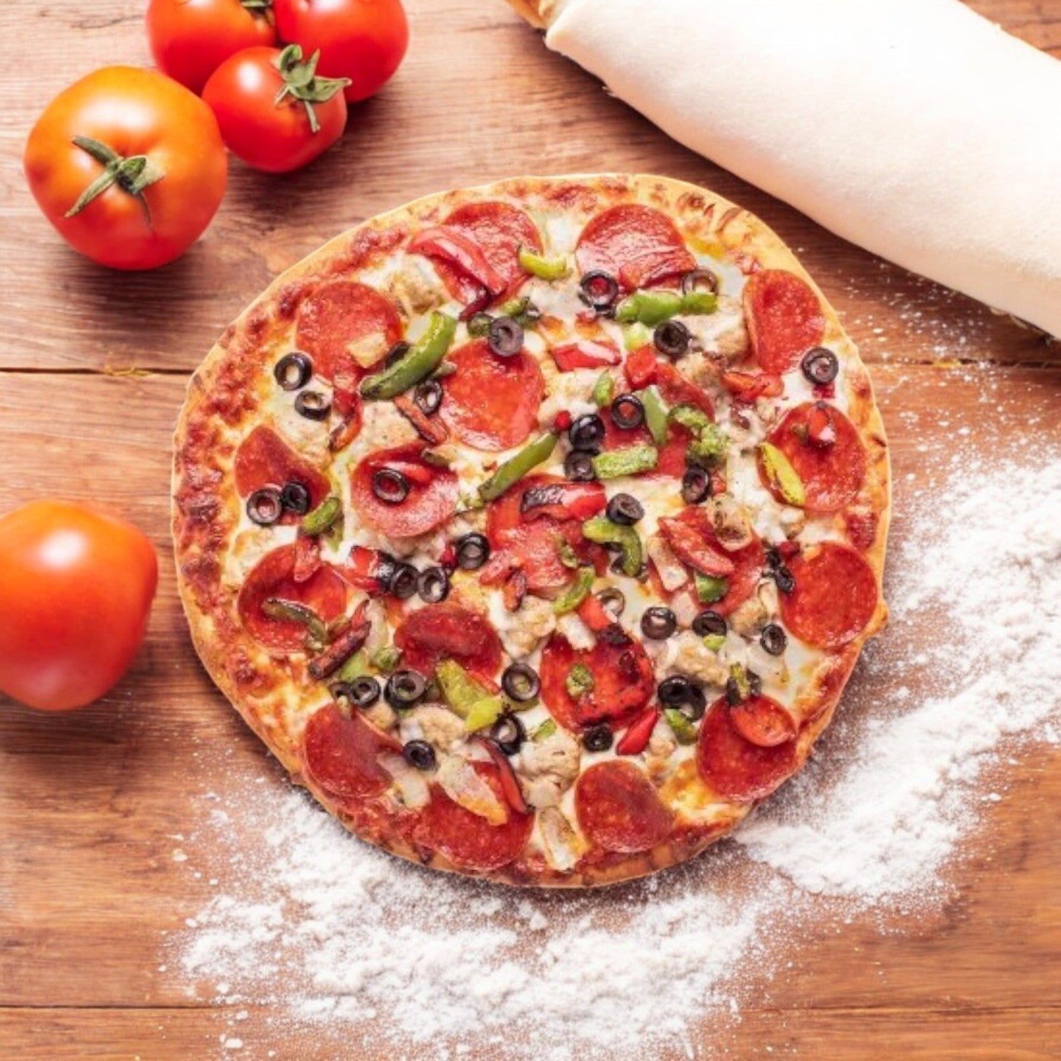 The Screamin Sicilian Supremus Maximus Pizza, 25 oz, features delicious pepperoni, olives, green peppers, and a cheesy blend on a wooden surface. Nearby are ripe tomatoes, a rolling pin, and scattered flour—a tasty pizza delight with every bite.