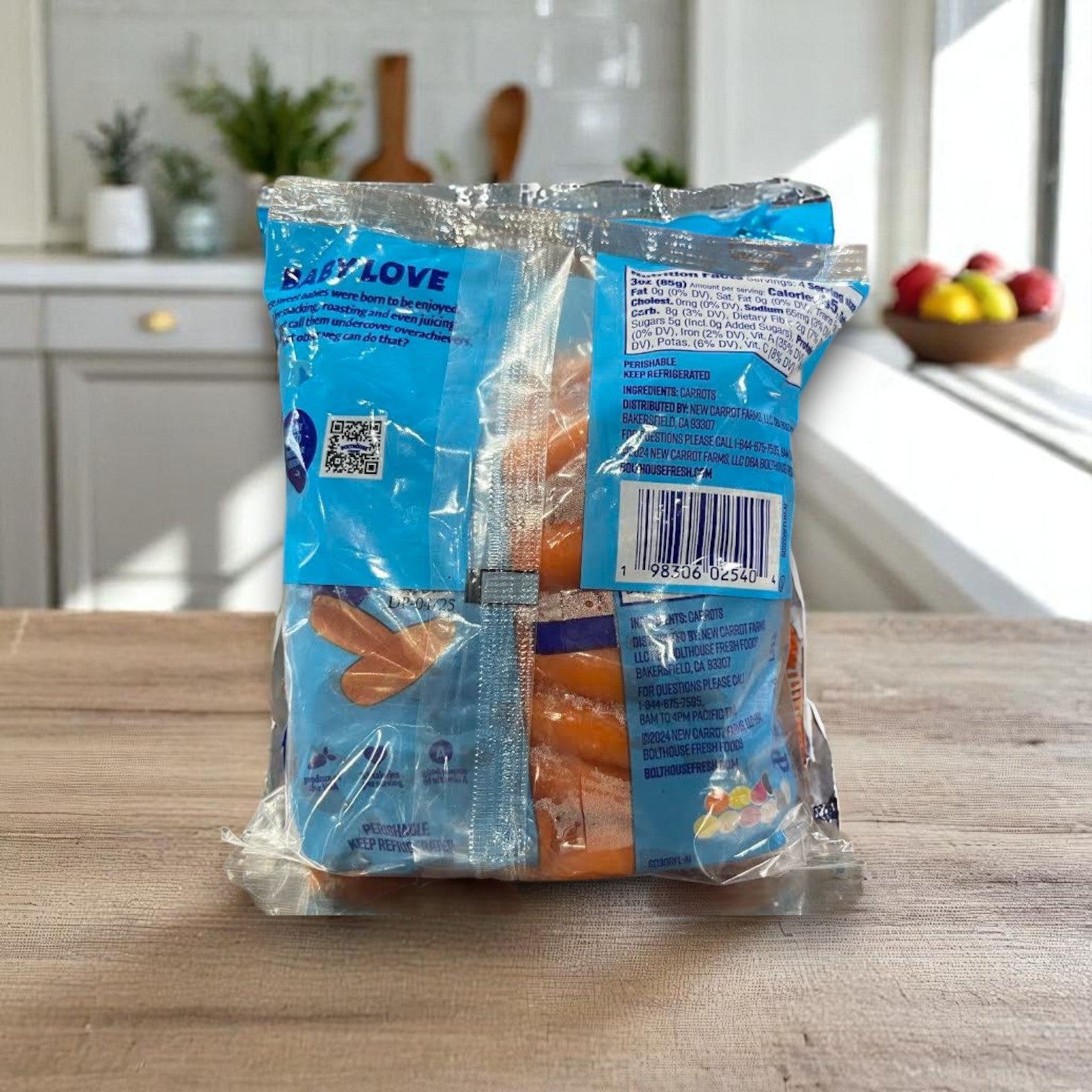 A 4-pack of Bolthouse Farms Carrots sits on a wooden counter in a bright kitchen, with fruit on the windowsill and cutting boards nearby—perfect for healthy snacking.