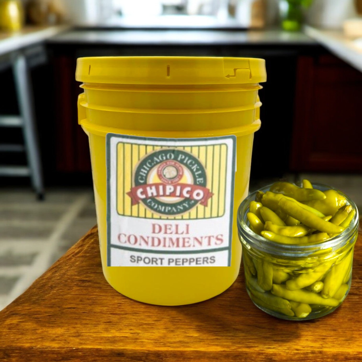 A 5-gallon yellow bucket of Vienna Beef Chipico Sport Peppers, ideal for foodservice or Chicago-style hot dogs, sits on the counter beside a small glass jar of green peppers.
