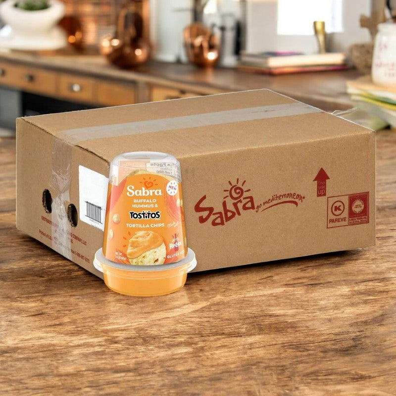 A box of Sabra Hummus Dip Buffalo With Tostitos, 2.4 oz., sits on a wooden table next to a large shipping case displaying the Sabra logo.