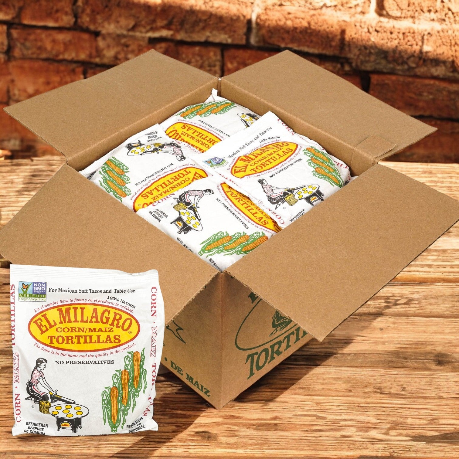 An open cardboard box with packs of El Milagro Yellow Corn Tortillas - 6" (40 packs of 1 dozen) features one pack at the front. These tortillas deliver authentic Mexican flavor and versatile size, set against a rustic brick wall backdrop.