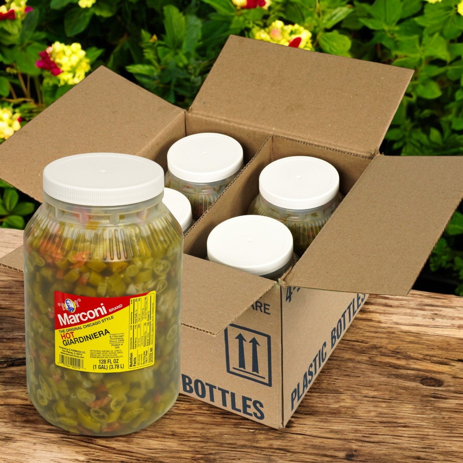 A jar of Marconi Giardiniera Hot from Marconi Foods is placed next to an open box labeled "Plastic Bottles," containing five more jars. The colorful pickled vegetables rest on wood with lush plants as a green backdrop.
