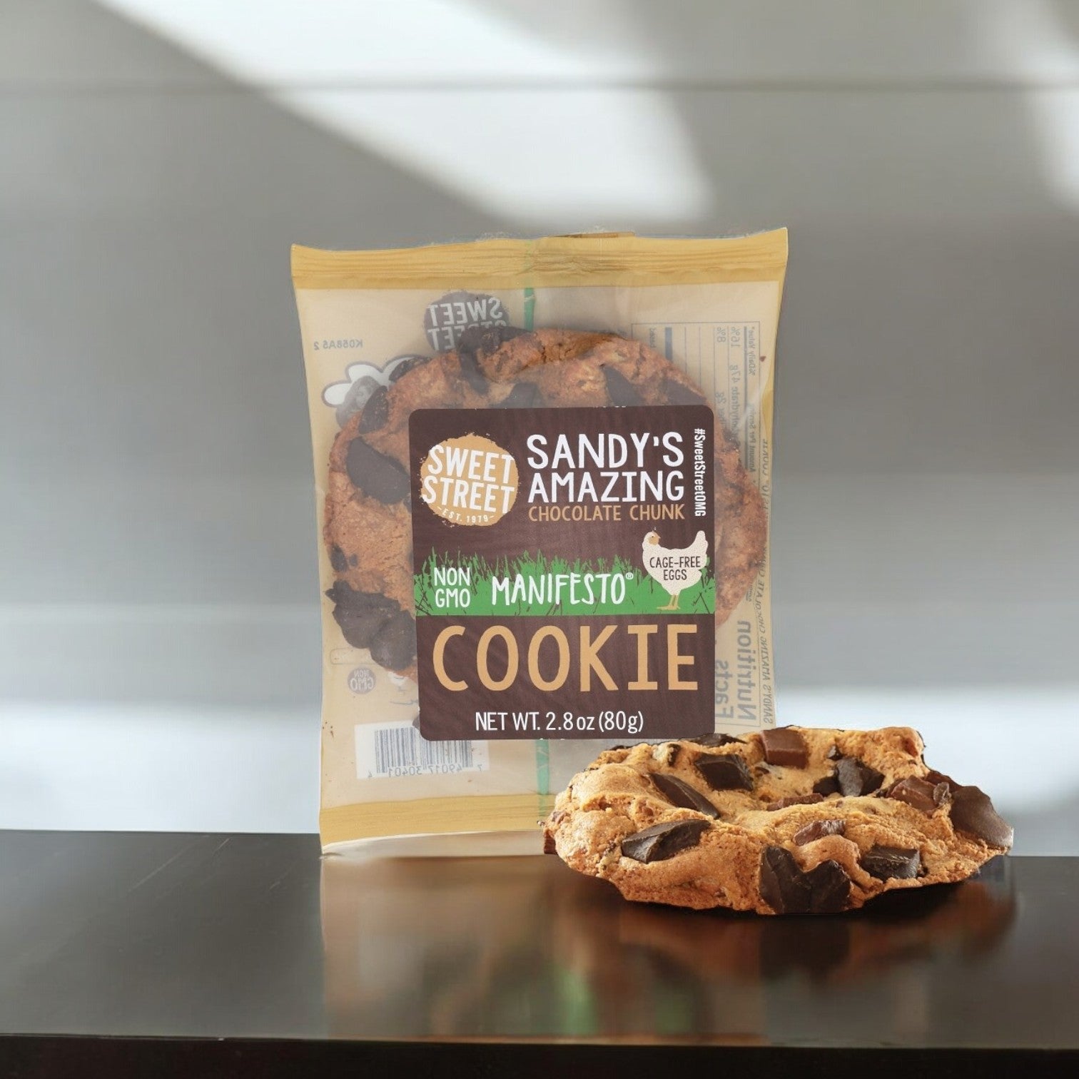 A large chocolate chunk cookie from Sweet Street sits beside its packaging on a dark surface. The package reads "Sandy's Amazing Chocolate Chunk Manifesto Cookies," showcasing these non-GMO cookies as the premium dessert choice.