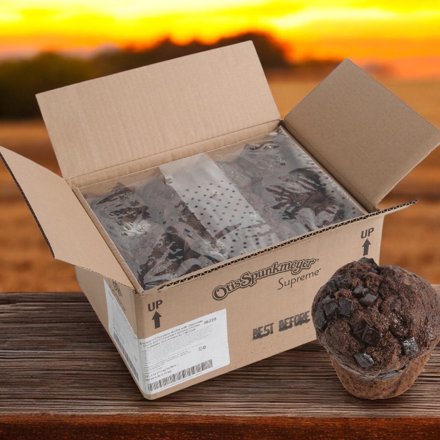 A cardboard box containing Otis Spunkmeyer Muffin Chocolate Chunk Supreme 24/4 oz. - Bulk Pack reveals its individually wrapped treats. On a wooden surface, one chocolate muffin entices from beside the box, with the sun setting over a tranquil field behind it.