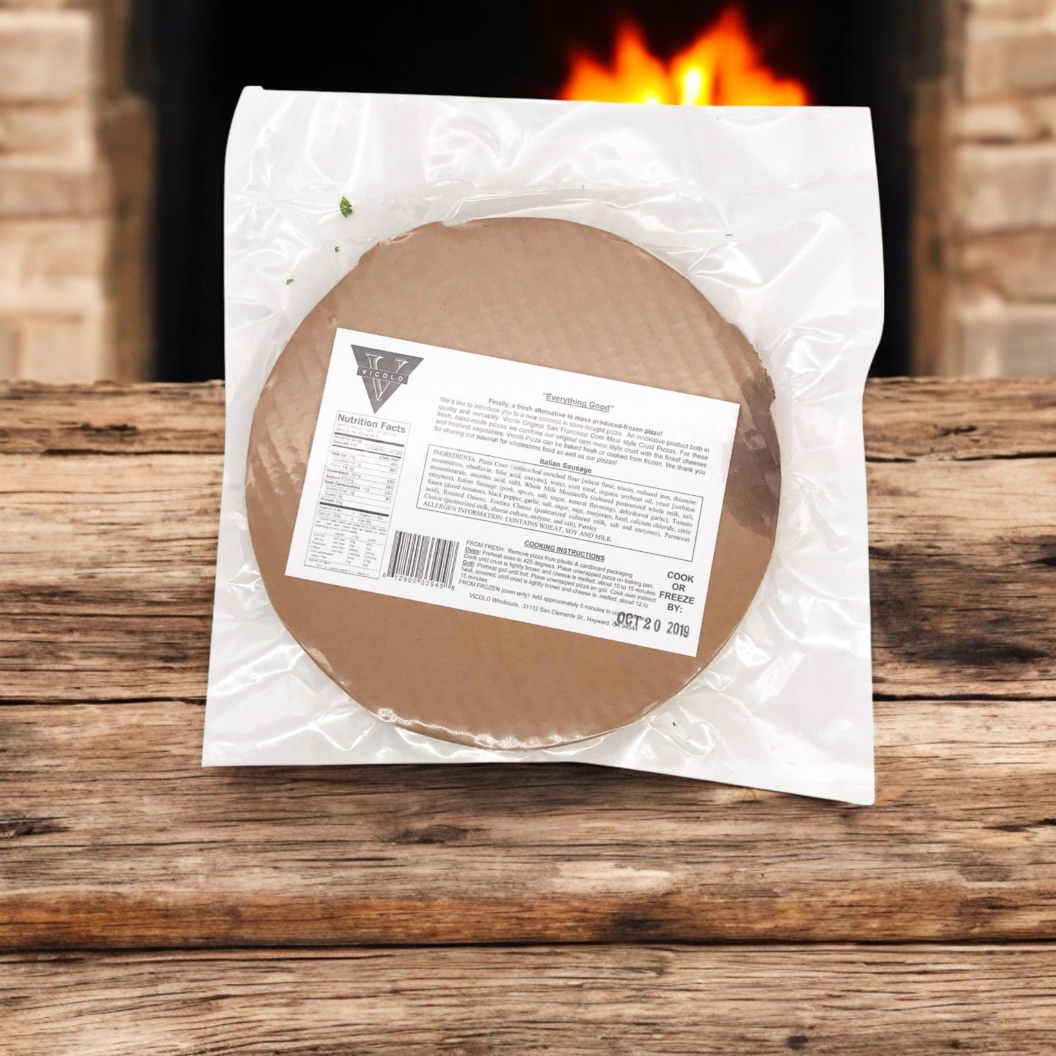 A sealed round watermelon soap package sits on wood, echoing the artistry of an Italian chef with Vicolo Itallian Sausage Pizza. The warm fireplace in the background resembles a wood-fired oven, enhancing the cozy ambiance.