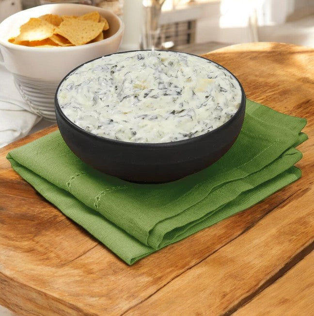 A black bowl of Member's Mark Spinach Artichoke Dip rests on a green napkin on a wooden surface, ideal for any party. In the background, tortilla chips are ready to pair with the creamy dip.