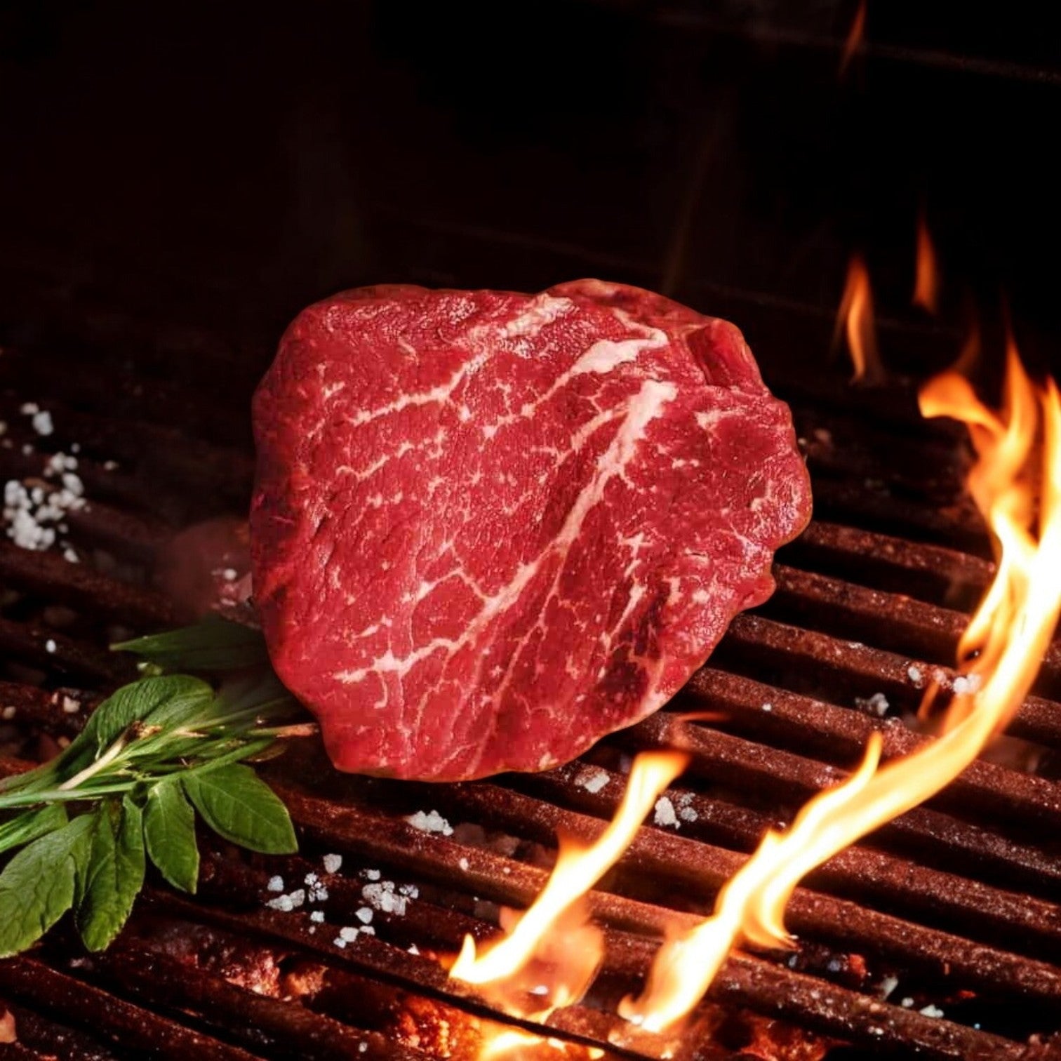 A 6oz Mino Prime Steaks filet sizzles on a hot grill, flames visible, adorned with fresh herbs and coarse salt scattered across the prime-grade beef.