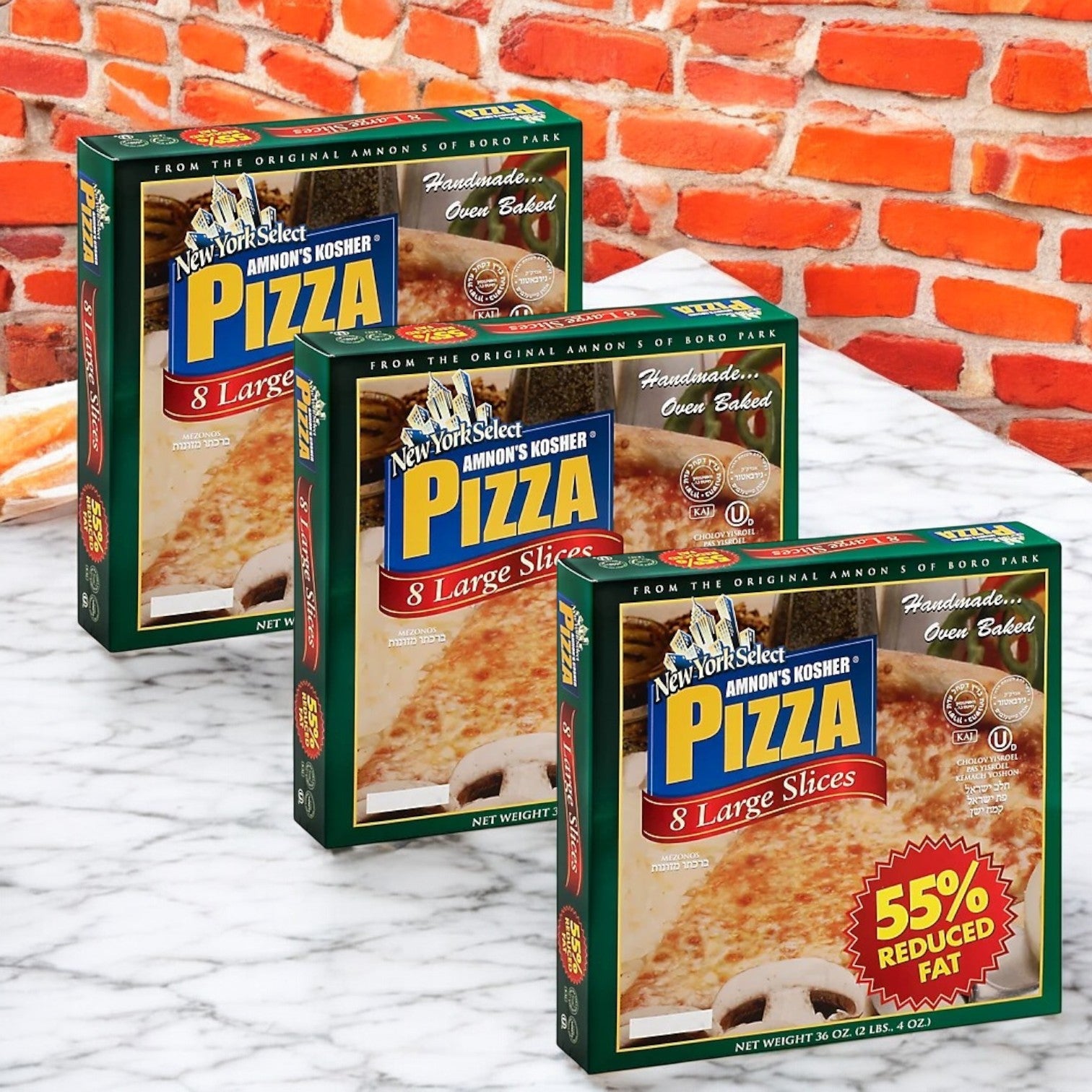 Displayed against a red brick wall on a marble surface are three boxes of Amnon's Kosher Pizza, 55% Reduced Fat Frozen 36 oz.- 3 Count, each with eight large slices. Savor this healthier pizza option offering delicious taste without the fat.