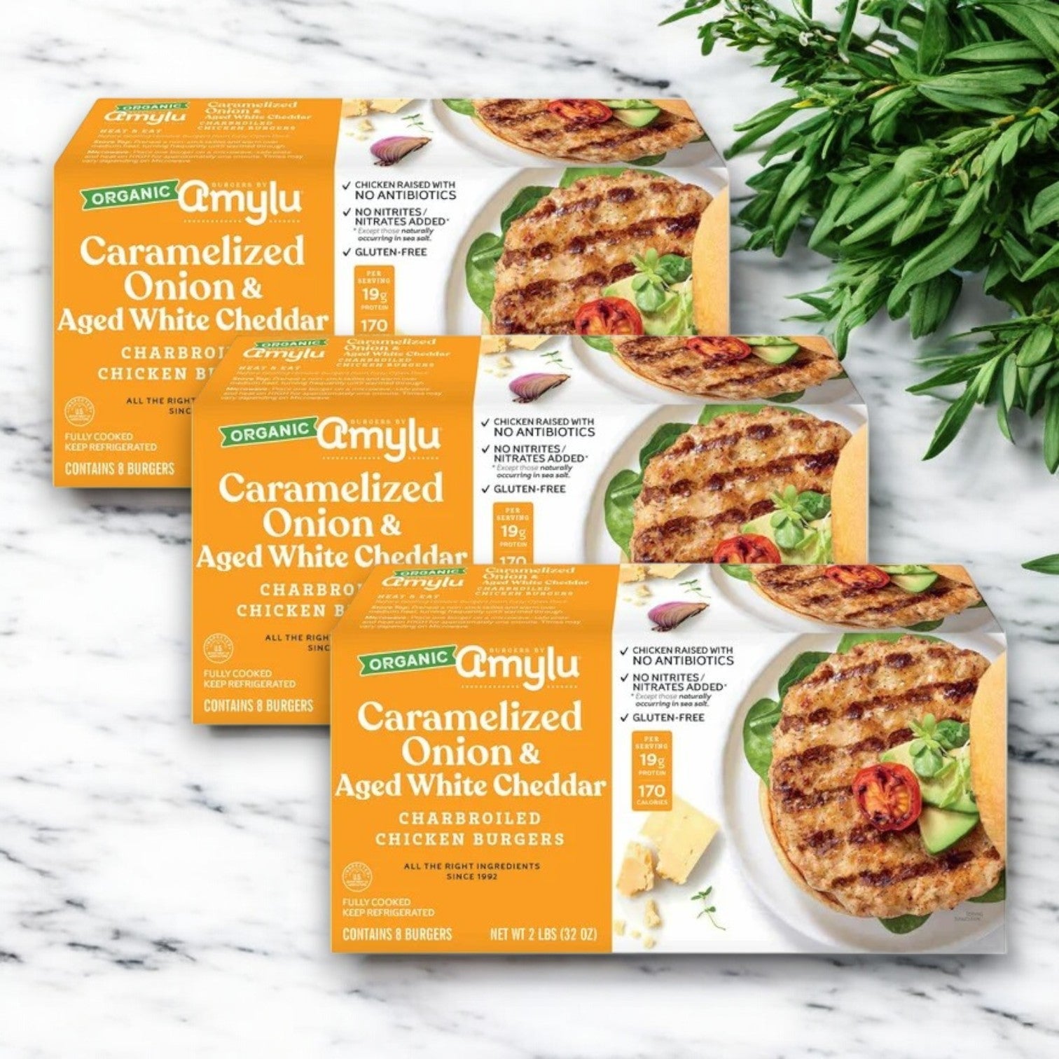 Three packages of Amylu Organic Chicken Burger with Caramelized Onion & Aged White Cheddar (32 oz, 24-Count) sit on a marble surface beside a small bunch of green herbs.