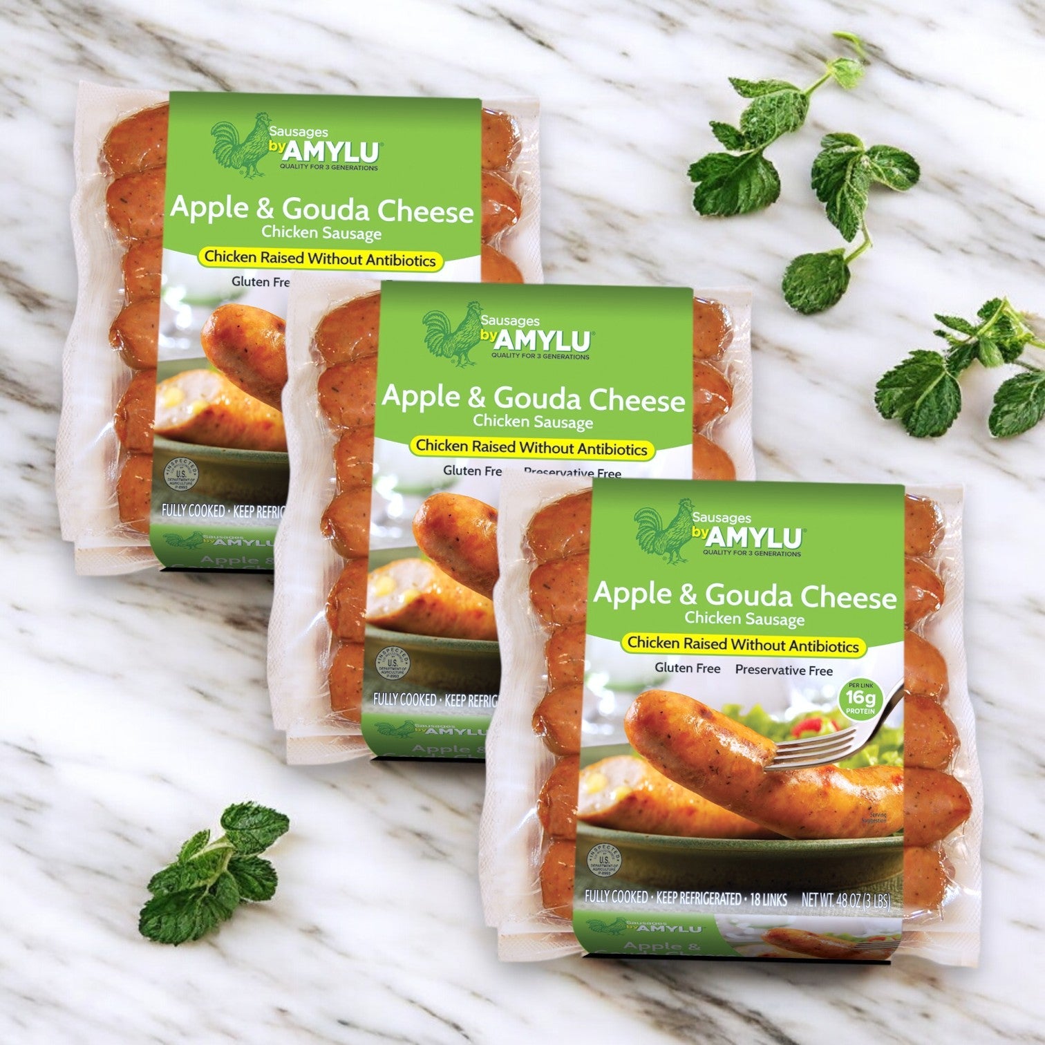Three packages of Amylu Apple & Gouda Cheese Chicken Sausage – 9 lbs (54 ct) are elegantly arranged on a marble surface with mint leaves. The packaging emphasizes their antibiotic-free and gluten-free features, offering a gourmet flavor in this convenient bulk pack.