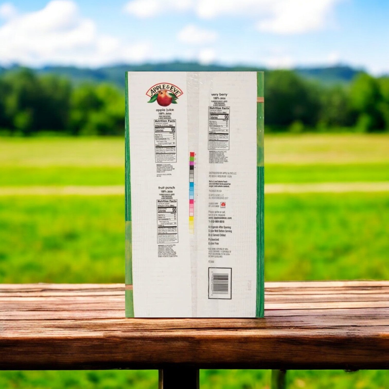On a wooden table outside, a box from the Apple & Eve 100% Juice Variety Pack by Refreshing Apple & Eve Juice displays nutritional info and logo against a grassy landscape.