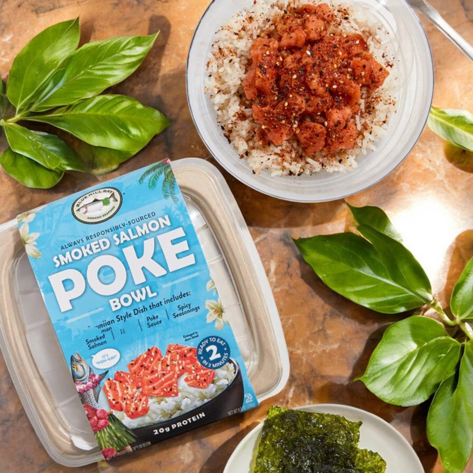 The Blue Hill Bay Smoked Salmon Poke Bowl, a 9.3 oz bundle of two gourmet seafood meals, features smoked salmon atop a rice bowl with seasoning, seaweed, and greens on a marble surface—convenience delivered to your door!.
