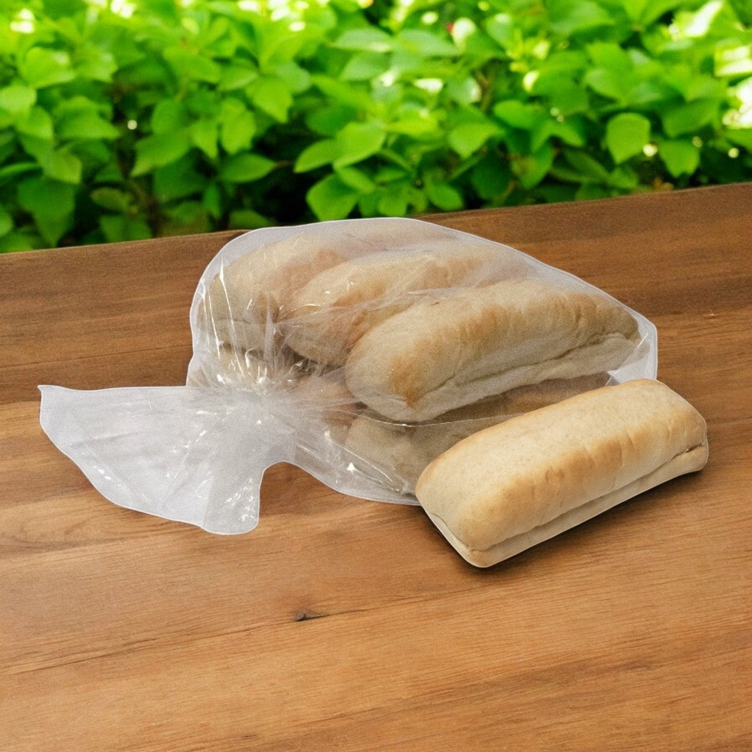 A partially opened plastic bag displays several Baker's Source Classic Italian Beef Hoagie Buns- 6 Pack Hoagie Hearth Hinged 6" Buns on a wooden surface, with green leaves in the background.