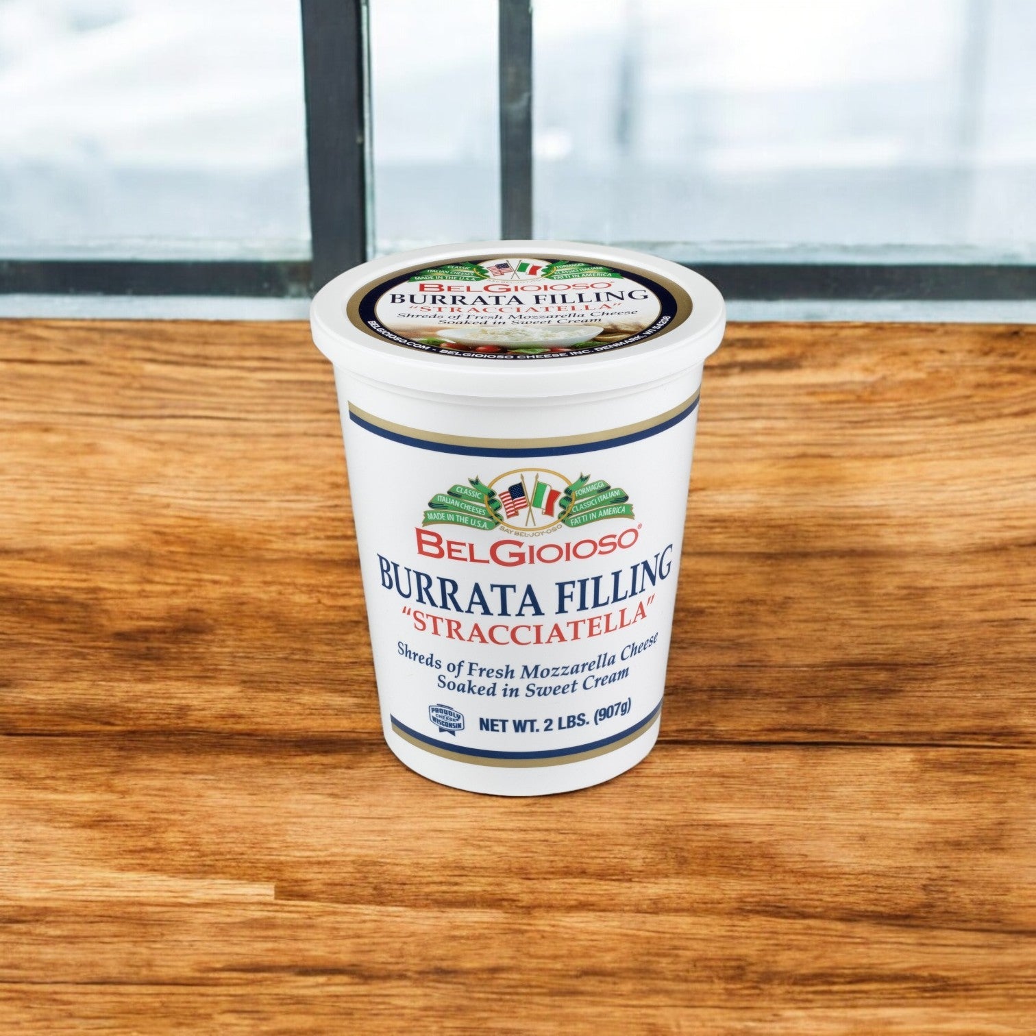 A BelGioioso Mozzarella Stracciatella Cup sits on a wooden surface by the window, showcasing its creamy texture.