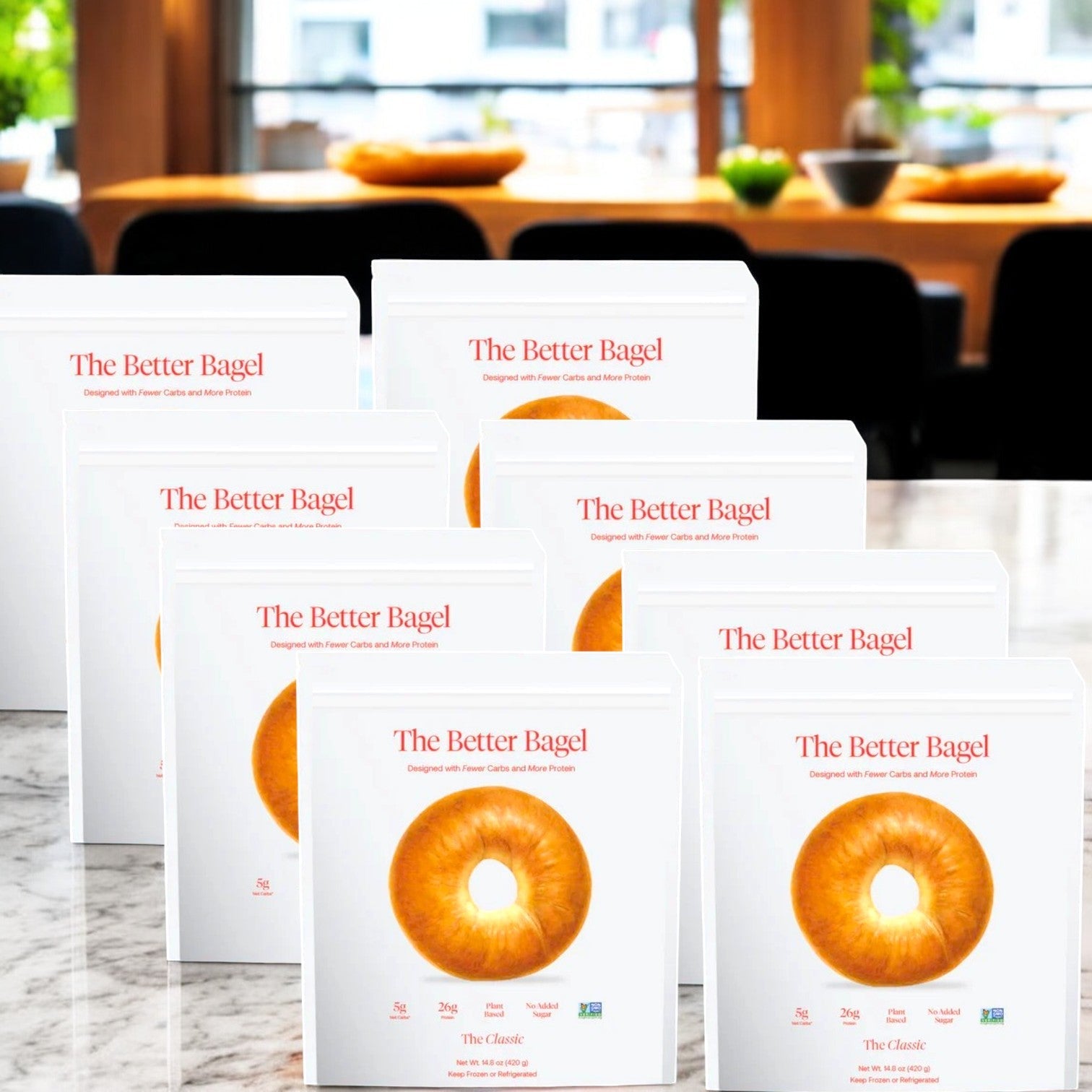 Eight bags of "BetterBrand - The Classic Better Bagel," known for their high-quality ingredients and irresistible soft, chewy texture, are displayed on a marble countertop in a room with large windows and a table in the background.