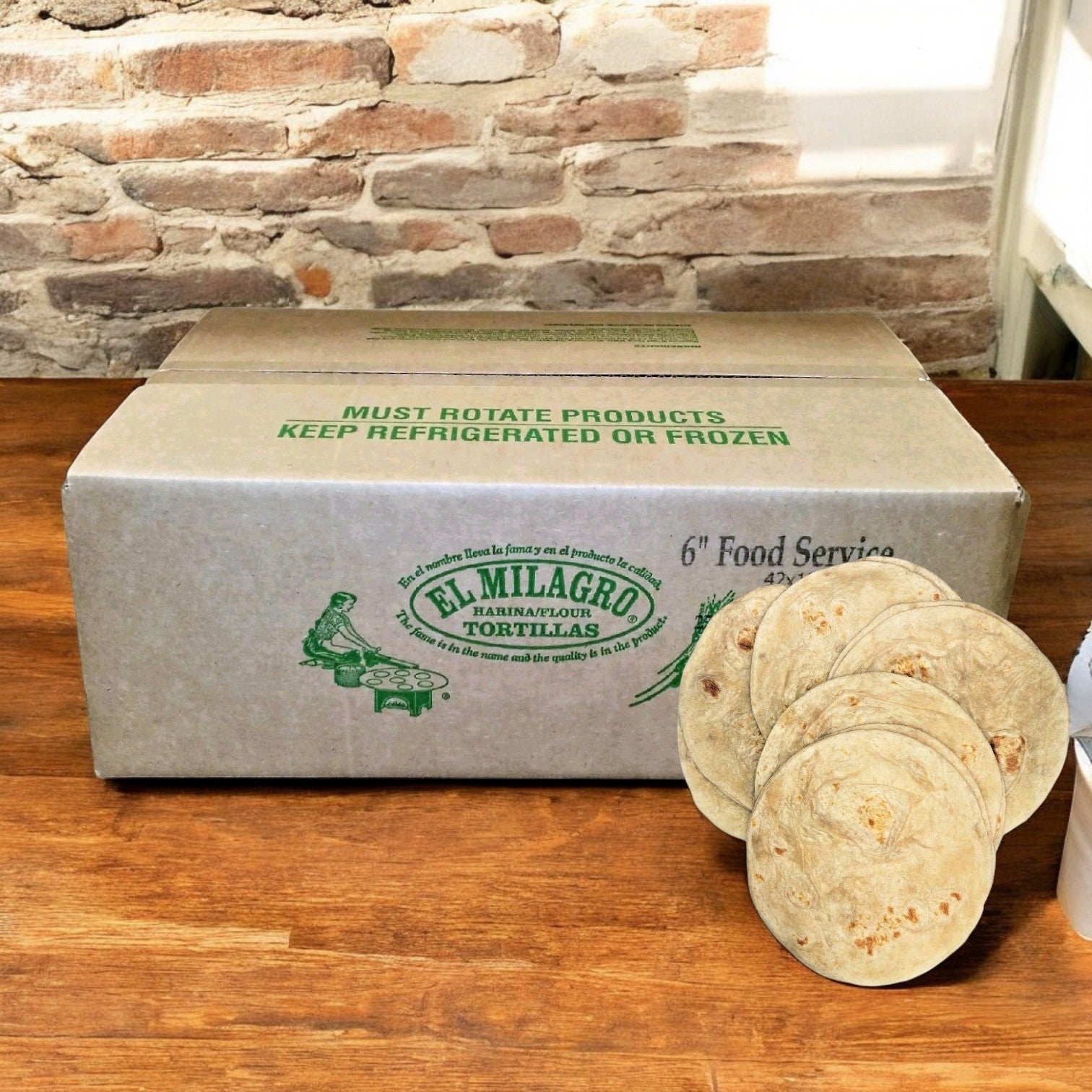 A cardboard box of El Milagro Flour Tortillas - 10-Inch - 12 Packs of 12 Dozen is displayed on a wooden table, highlighting their authentic flavor with stacks of tortillas arranged in front against a rustic brick wall.