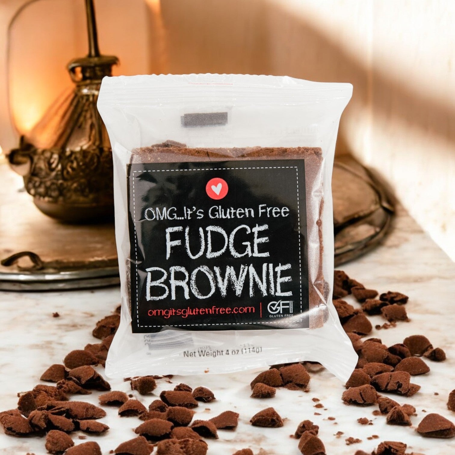 A packaged "OMG It's Gluten Free, Brownie Fudge Gluten Free- 1 Case, 24 Pack" rests on a marble surface with scattered fudge brownie crumbs.