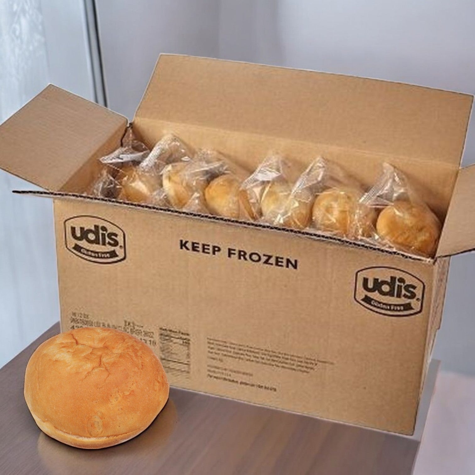 A cardboard box labeled "Udi's" contains individually wrapped, gluten-free hamburger buns and displays "Keep Frozen." One soft and sturdy bun rests outside the box.