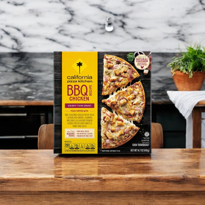 A box of California Pizza Kitchen BBQ Chicken Thin Crust - 1 Count rests invitingly on a wooden table in a cozy kitchen, offering a gourmet taste of California at home.