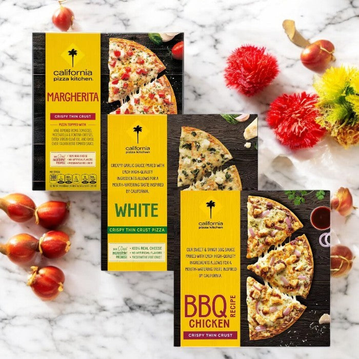Three California Pizza Kitchen thin crust pizzas—Margherita, White, and BBQ Chicken—are displayed on a marble surface with decorative red and yellow flowers.